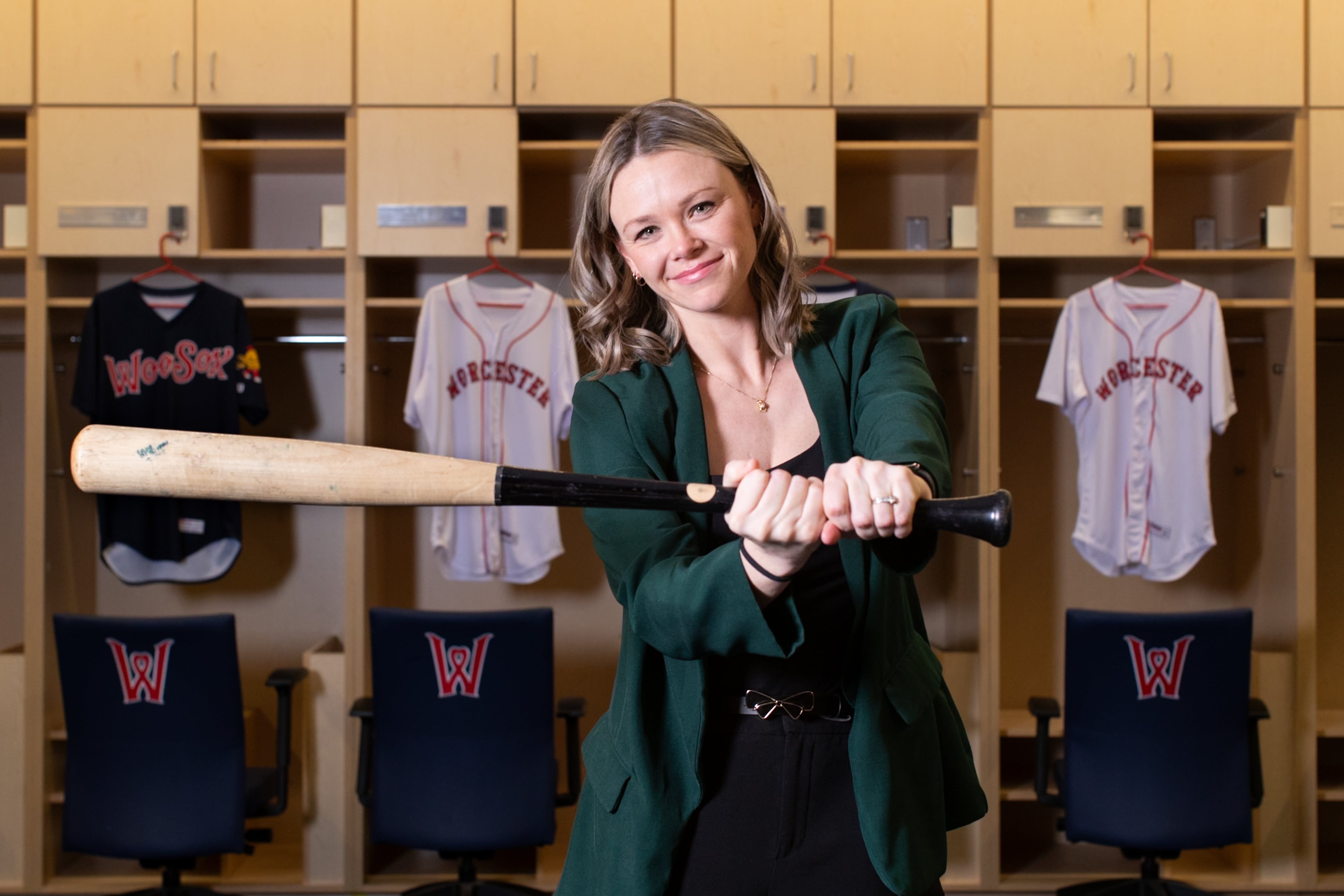 A woman wearing a black outfit and green jacket swings a baseball bat in front of a panel of lockers with Worcester Red Sox jerseys hanging in them.