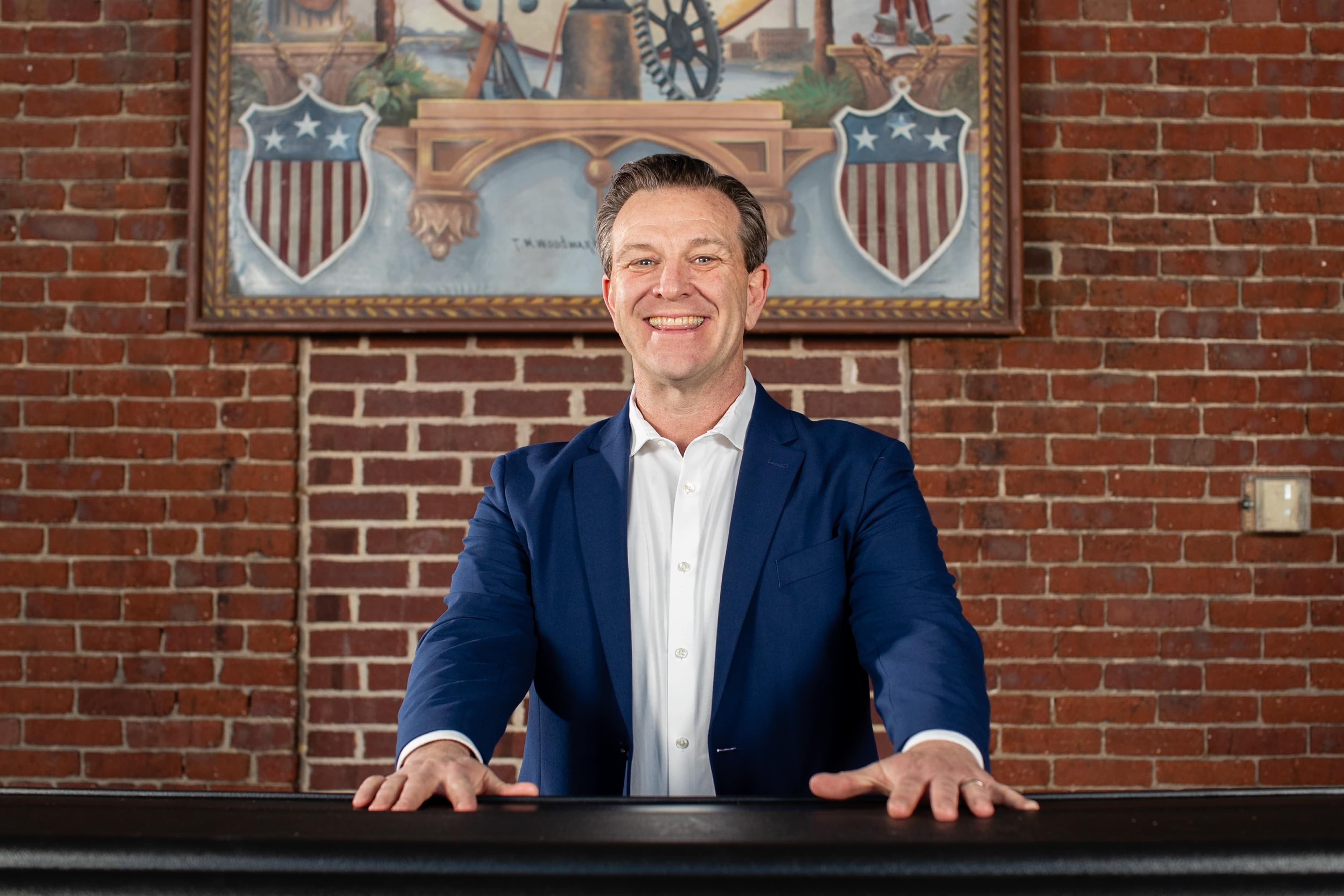 A man in a blue blazer stands in front of a brick wall.