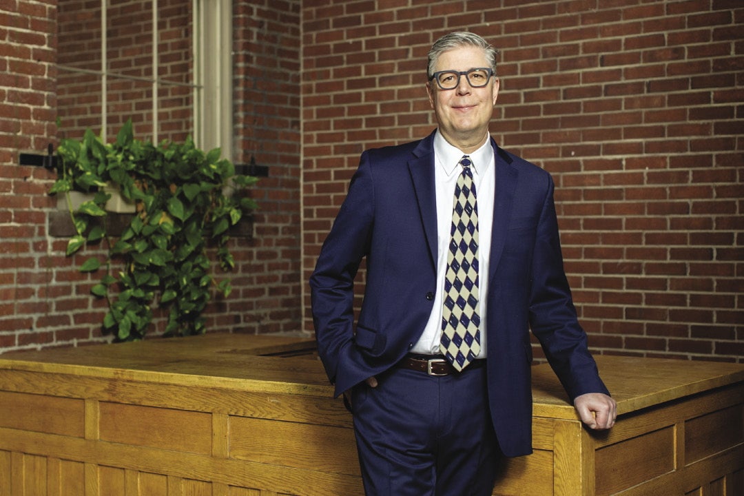 A man with grey hair wears grey glasses, a dark blue suit, white button down, and patterned tie while leaning on a wooden table top inside a brick building.