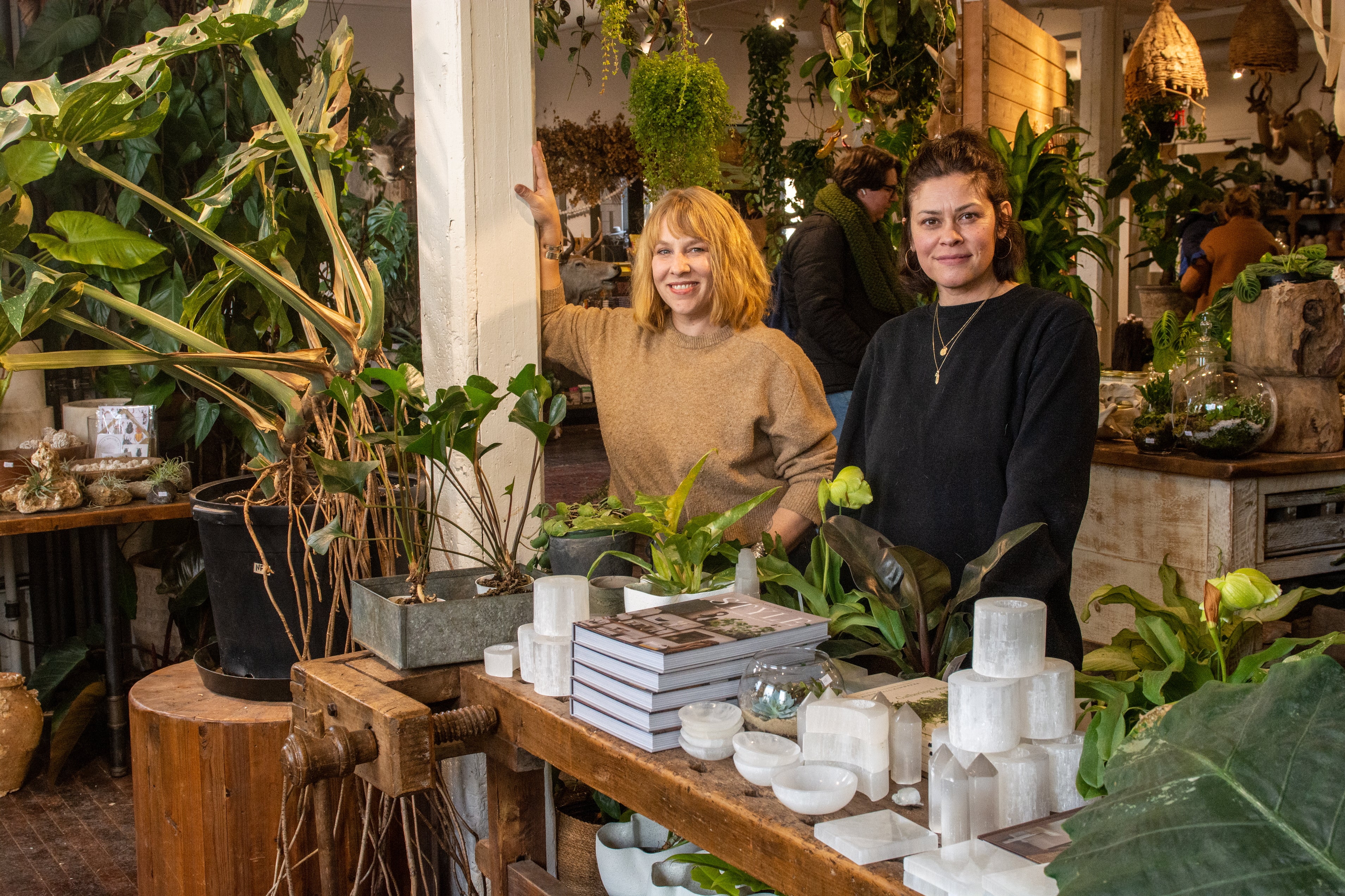 One woman with blonde hair and a woman with dark brown hair stand side by side in a store with house plants hanging from the ceiling and on tables with books, candle holders, and other home decor on display.