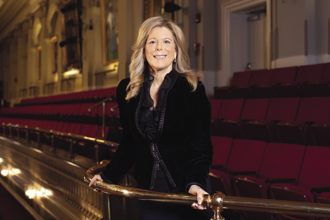 A woman with long blonde hair wears a black top while holding a railing in a theater auditorium.