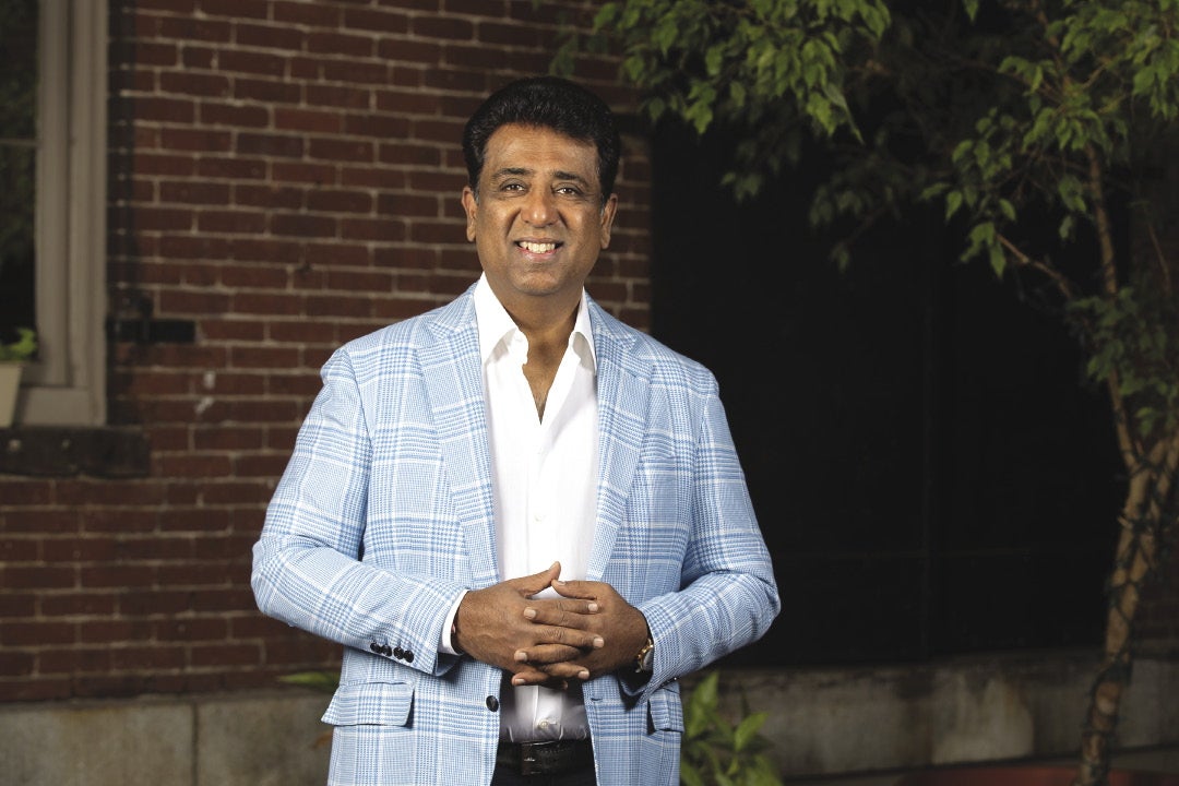 A man with short black hair wears a light blue plaid jacket, and a white button down while standing with his hands together in front of a brick building.