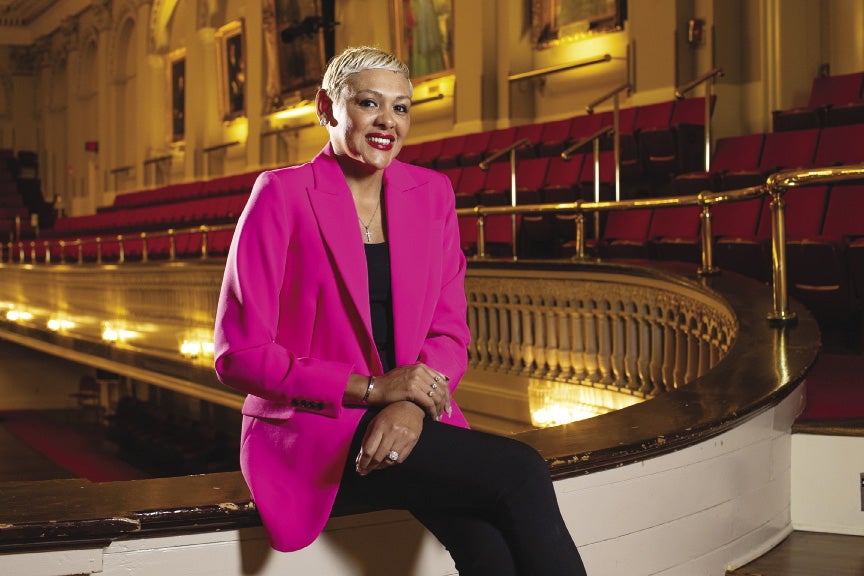 A woman with short blonde hair wears a neon pink suit jacket, black pants, and a black top while sitting in an auditorium.