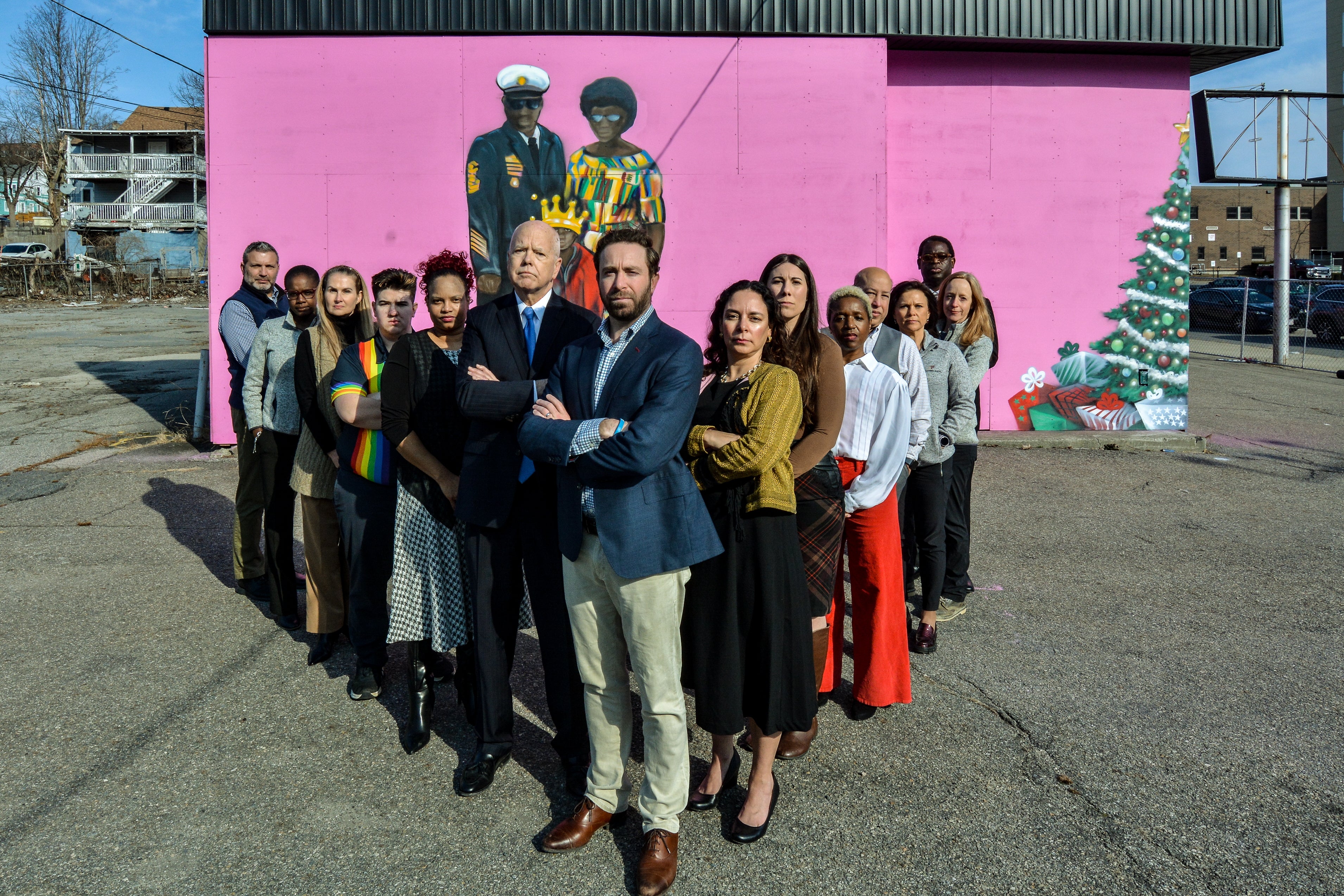 A group of people stand in a V formation in front of a pink mural outside.