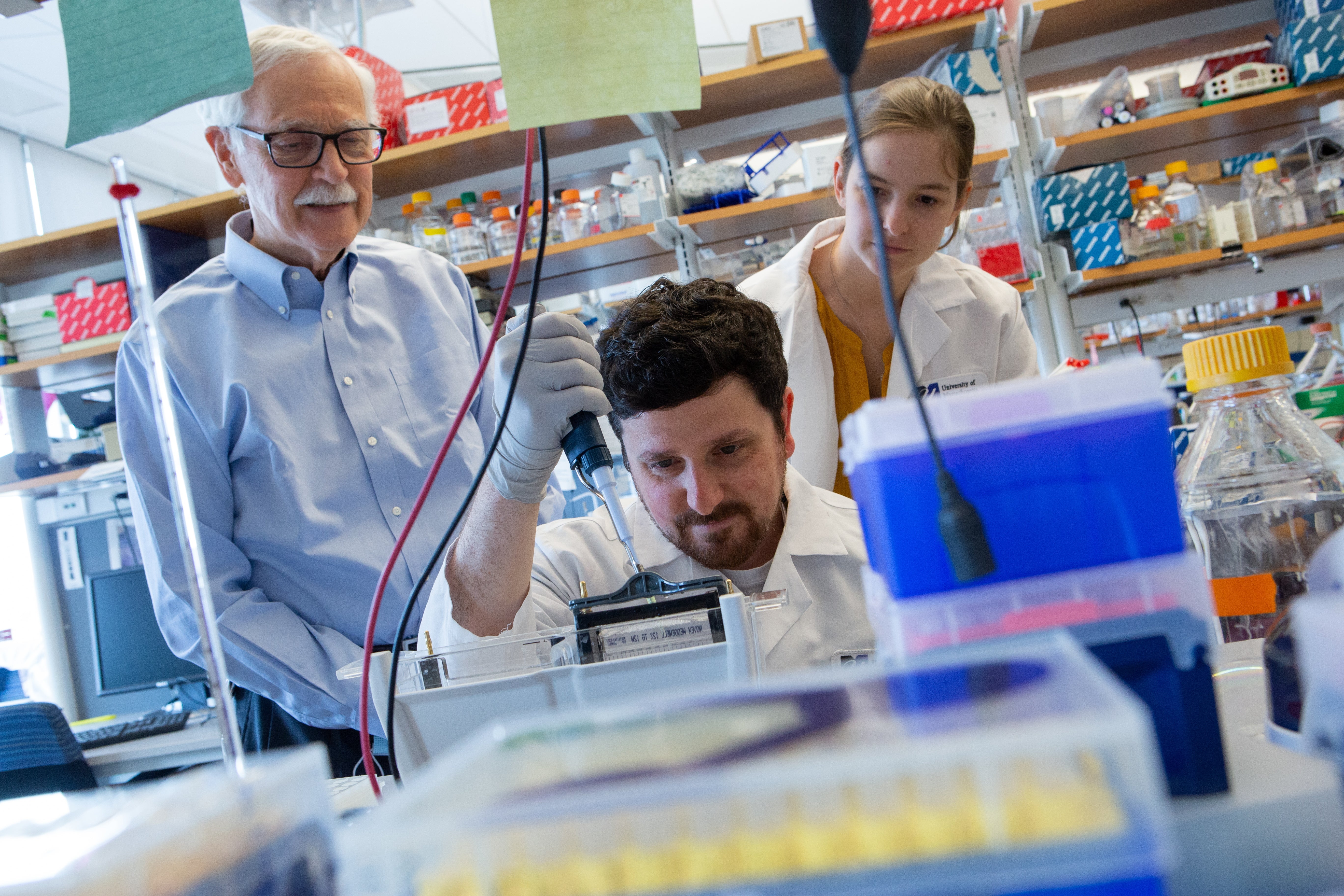 Two people stand behind a man sitting inside a room filled with research equipment.