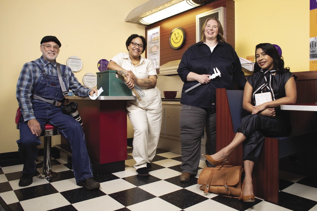 Three women and one man dressed in old-timey manufacturing outfits pose in a museum exhibit.