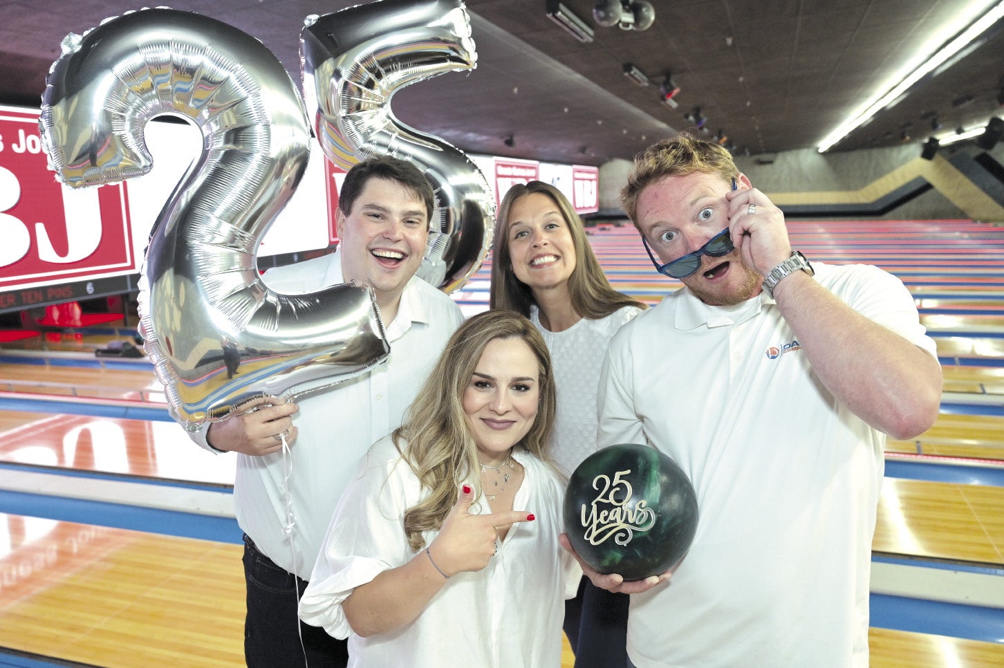 Four people in white shirts stand in a bowling alley, holding balloons and a bowling ball.