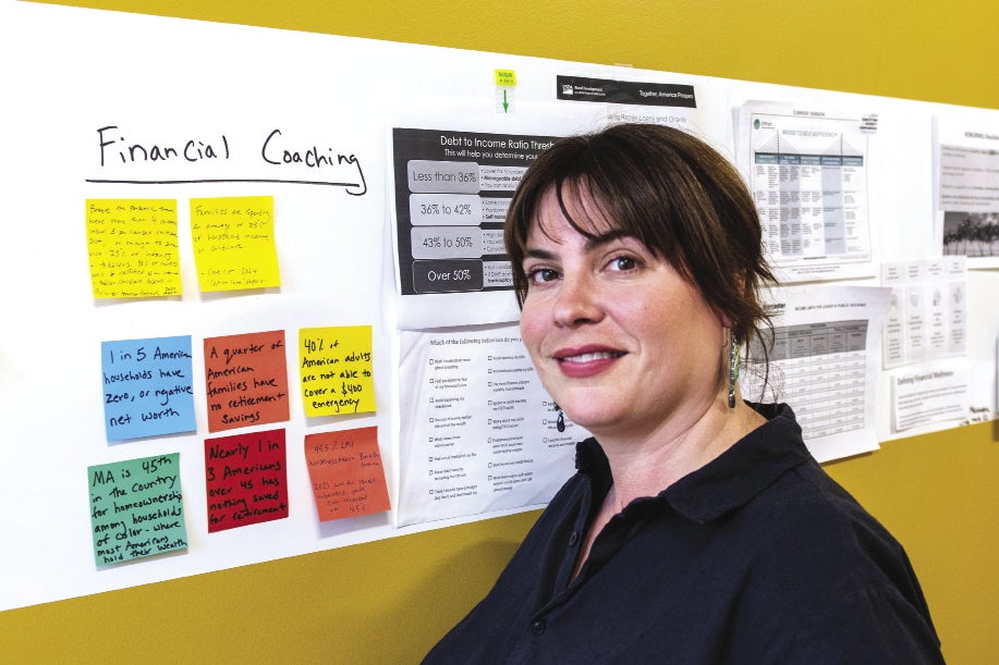 A woman with brown hair pulled back and bangs stands in front of a yellow wall with papers and sticky notes lined along it