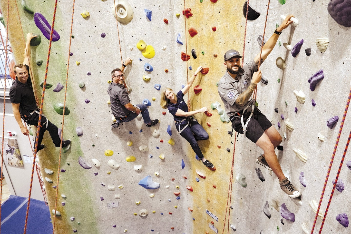 Three men and a woman hang from ropes on a climbing wall.