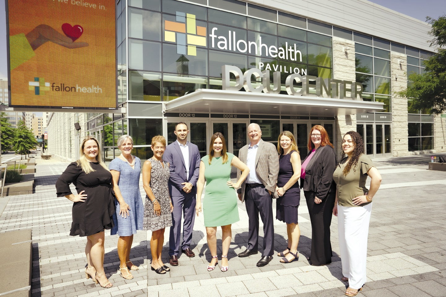 Seven women and two men stand in front of the DCU Center in Worcester on a sunny day.