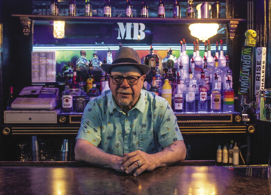A man leans forward against a bar wearing a light blue short sleeve button down, glasses and a fedora in front of shelves holding alcohol bottles.