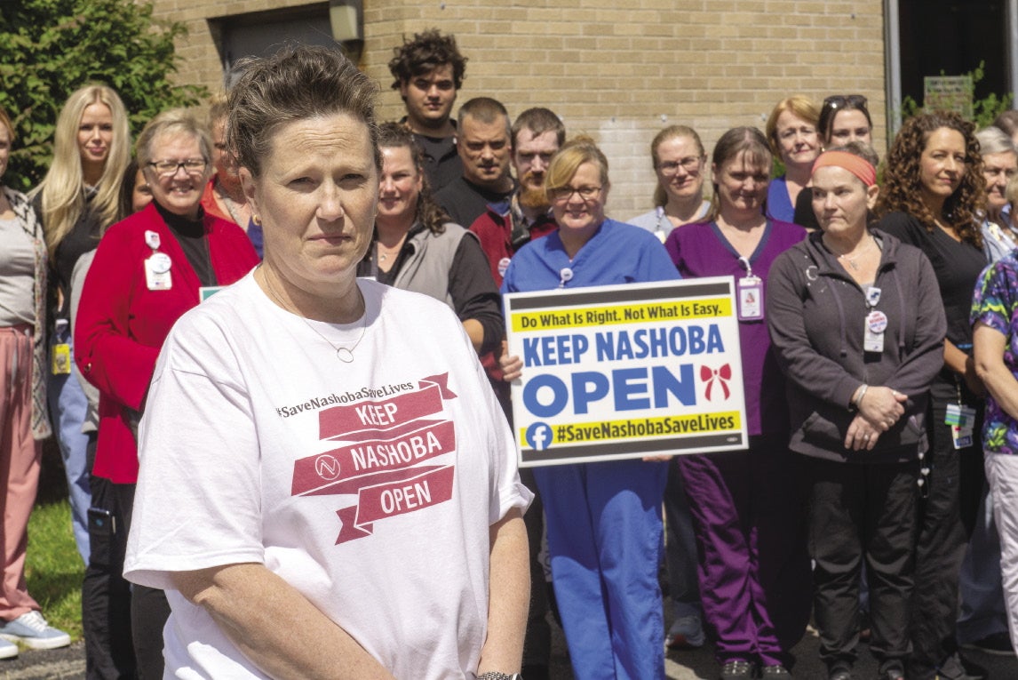 A woman with a white t-shirt stands in front of a group of hospital workers.