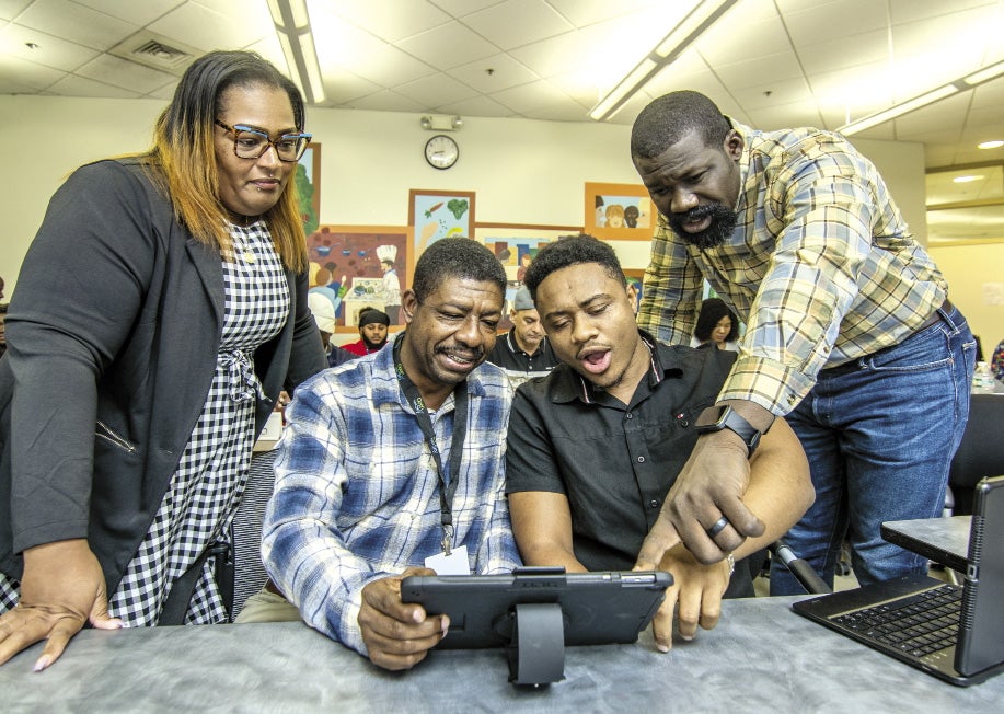 Two people stand looking over the outside shoulders of two men sitting next to each other: all are looking at an iPad and the two men on the right are pointing at it.