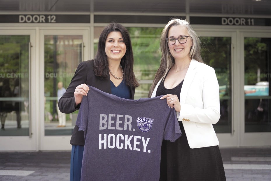 Two women stand in front of the DCU Center holding a jersey that says "Beer. Hockey."