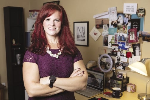 A woman with red hair and a purple shirt stands with har arms crossed in front of a desk with a laptop and photos on the wall above it.