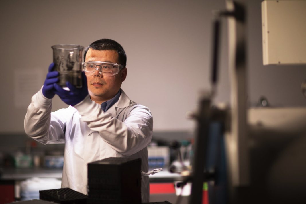 A man wearing a white lab coat and safety goggles holds up a beaker